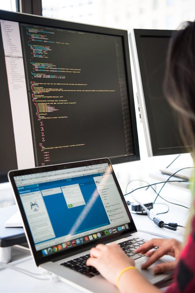 person-using-both-computer-and-a-laptop-1181673 Programmer working on code across multiple screens in a modern office setting.